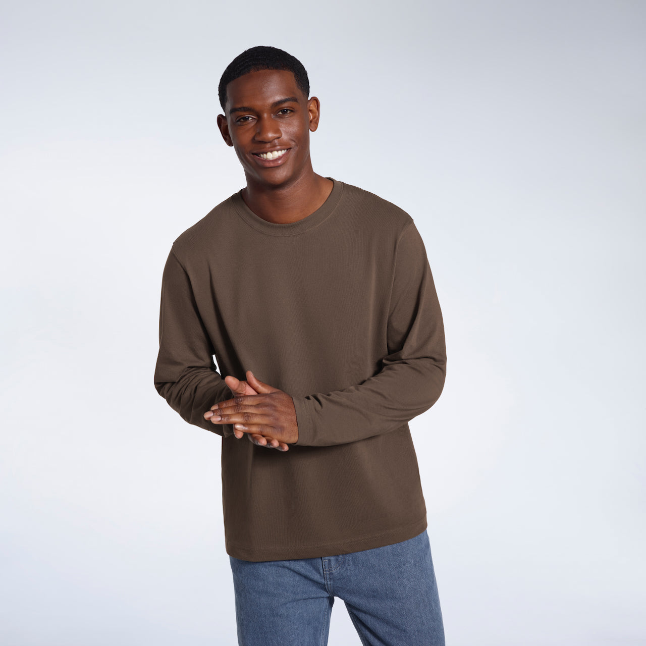 A man smiling into the camera with his hands together, modelling a plain dark khaki long sleeve organic cotton t shirt.