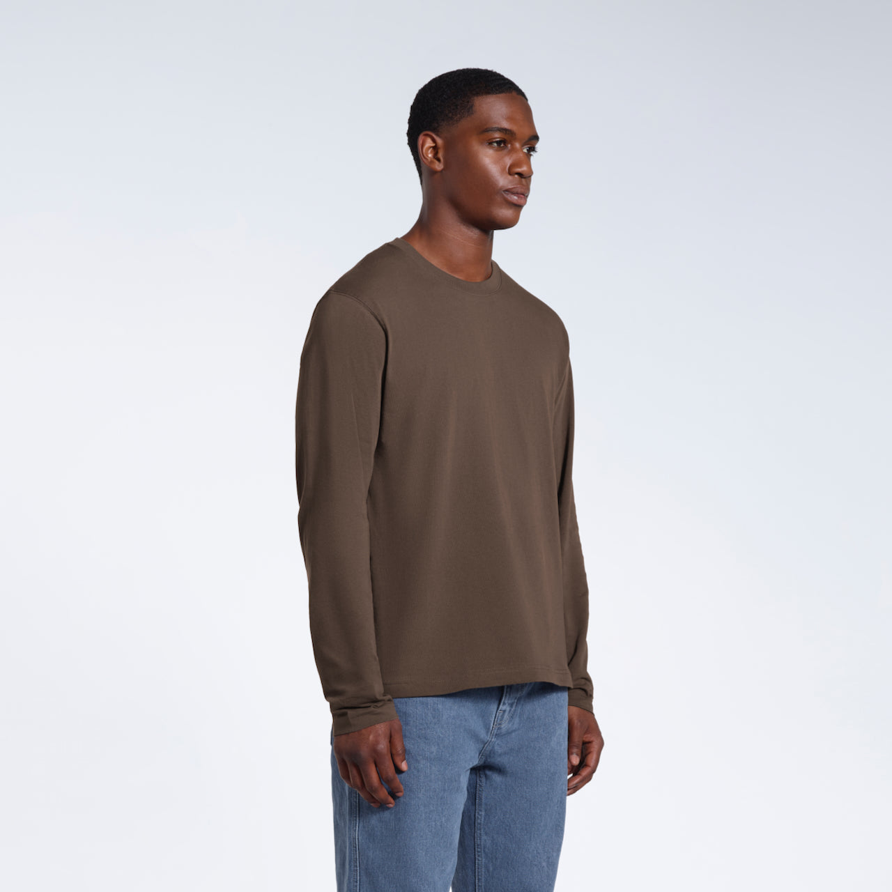 A man looking away from the camera with his arms by his sides, modelling a plain dark khaki long sleeve organic cotton t shirt.
