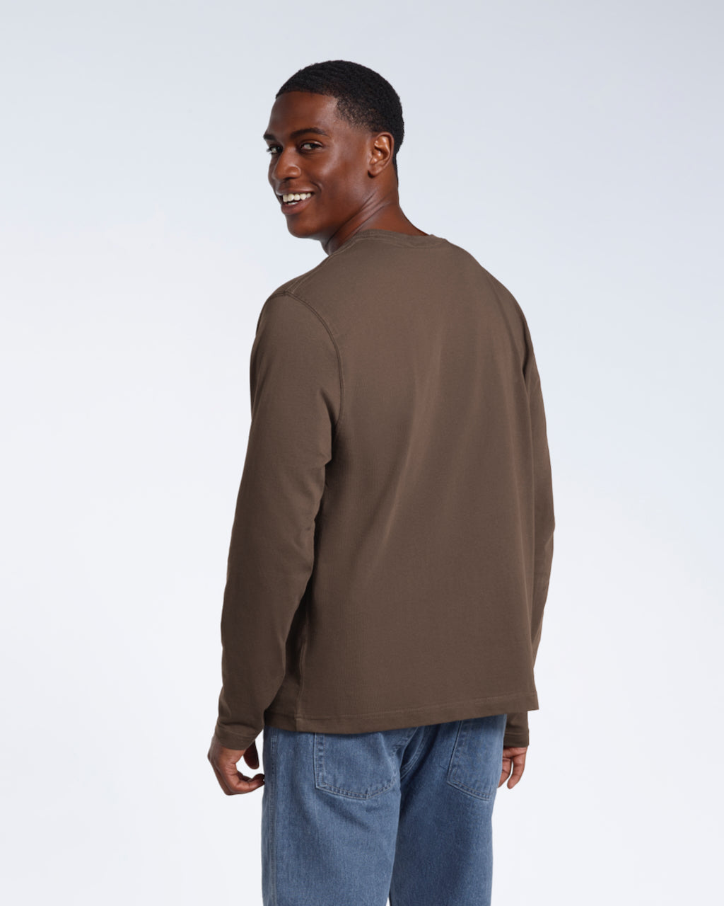 A man facing away from the camera looking back and smiling, modelling a plain dark khaki long sleeve organic cotton t shirt.