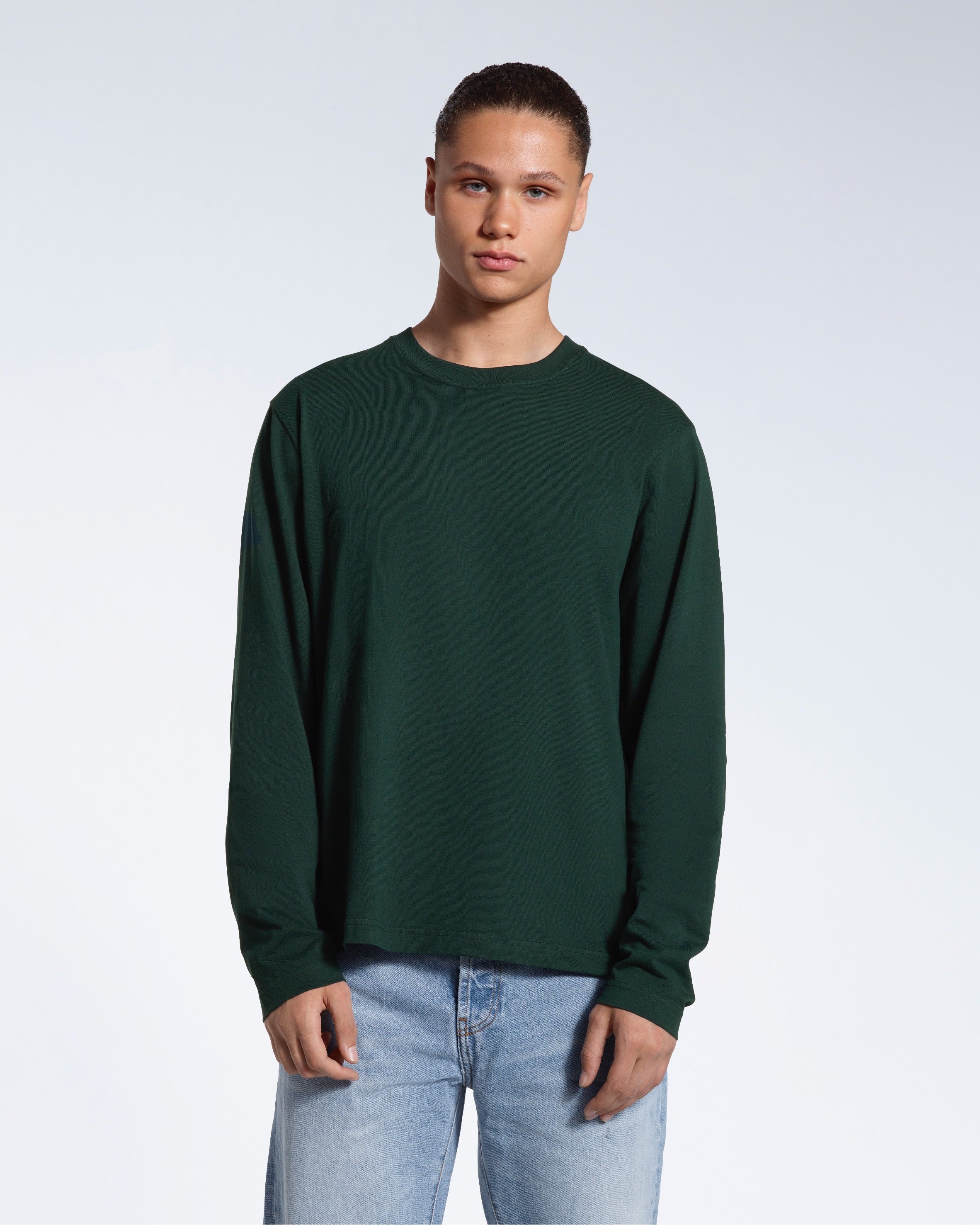 A man standing front on with his arms by his sides, modelling a plain green long sleeve organic cotton t shirt.