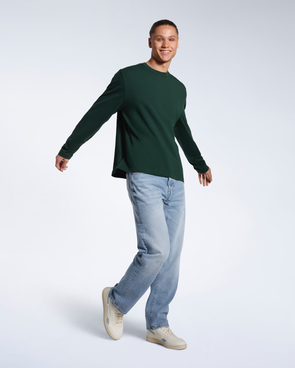 A man walking and smiling, modelling a plain green long sleeve organic cotton t shirt.