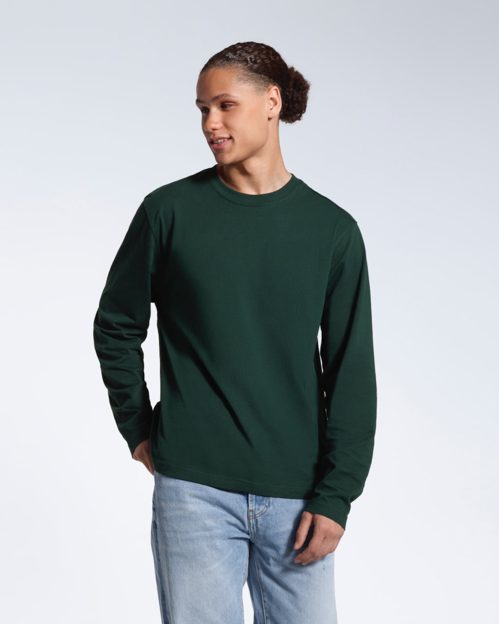 A man looking away from the camera with his hand in his back pocket, modelling a plain green long sleeve organic cotton t shirt.