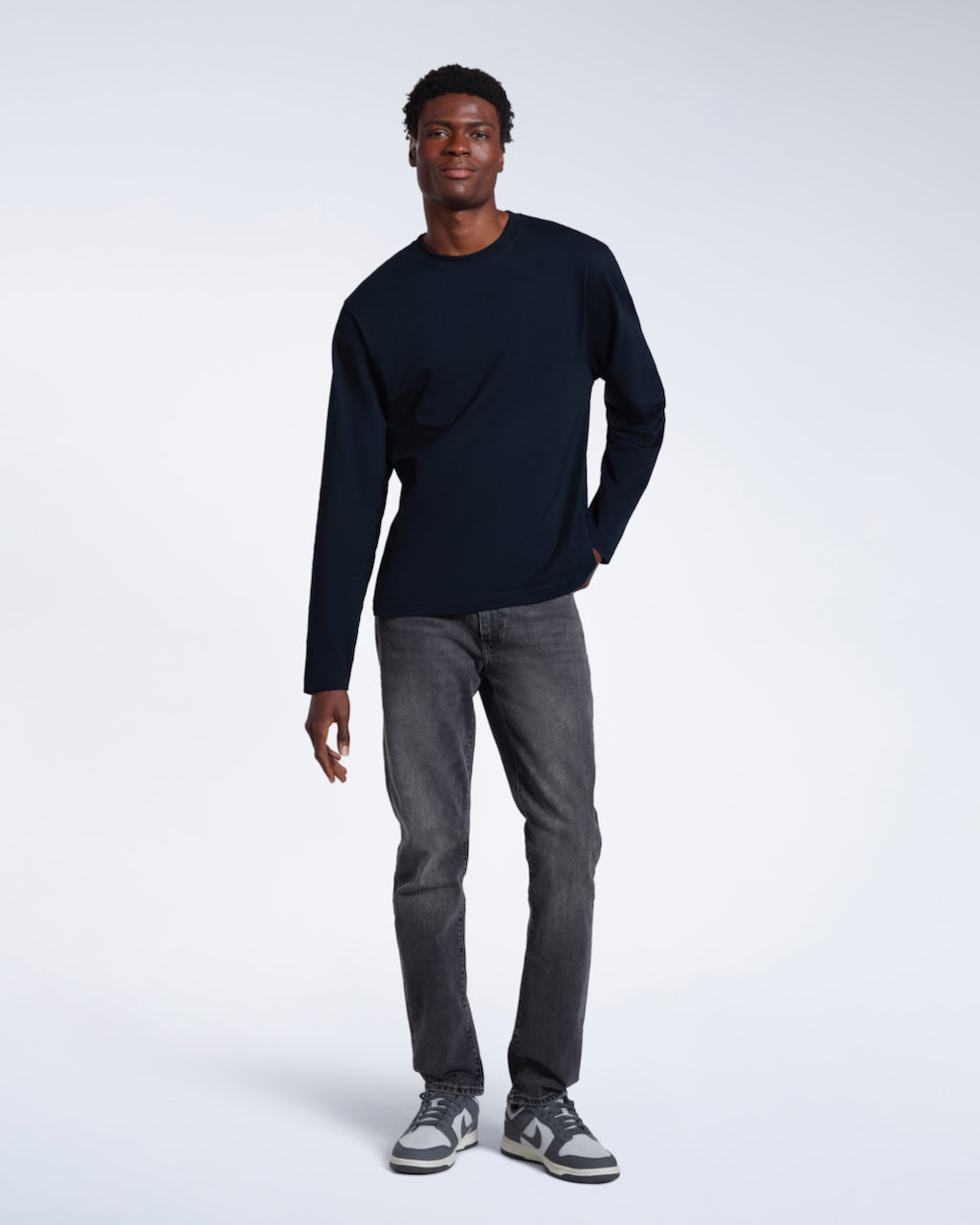 A full body photo of a man looking into the camera, modelling a plain navy long sleeve organic cotton t shirt.
