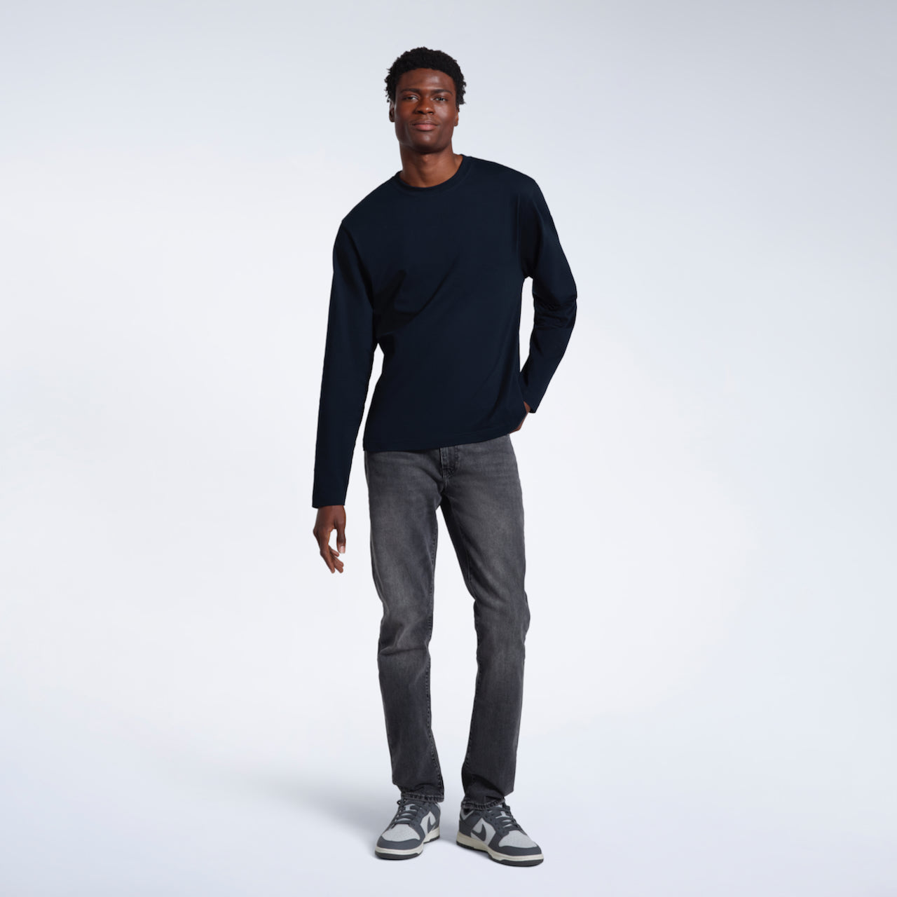 A full body photo of a man looking into the camera, modelling a plain navy long sleeve organic cotton t shirt.