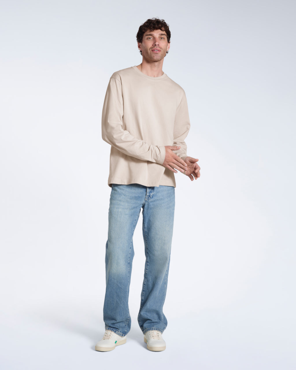 A full body photo of a man looking into the camera, modelling a plain stone long sleeve organic cotton t shirt.