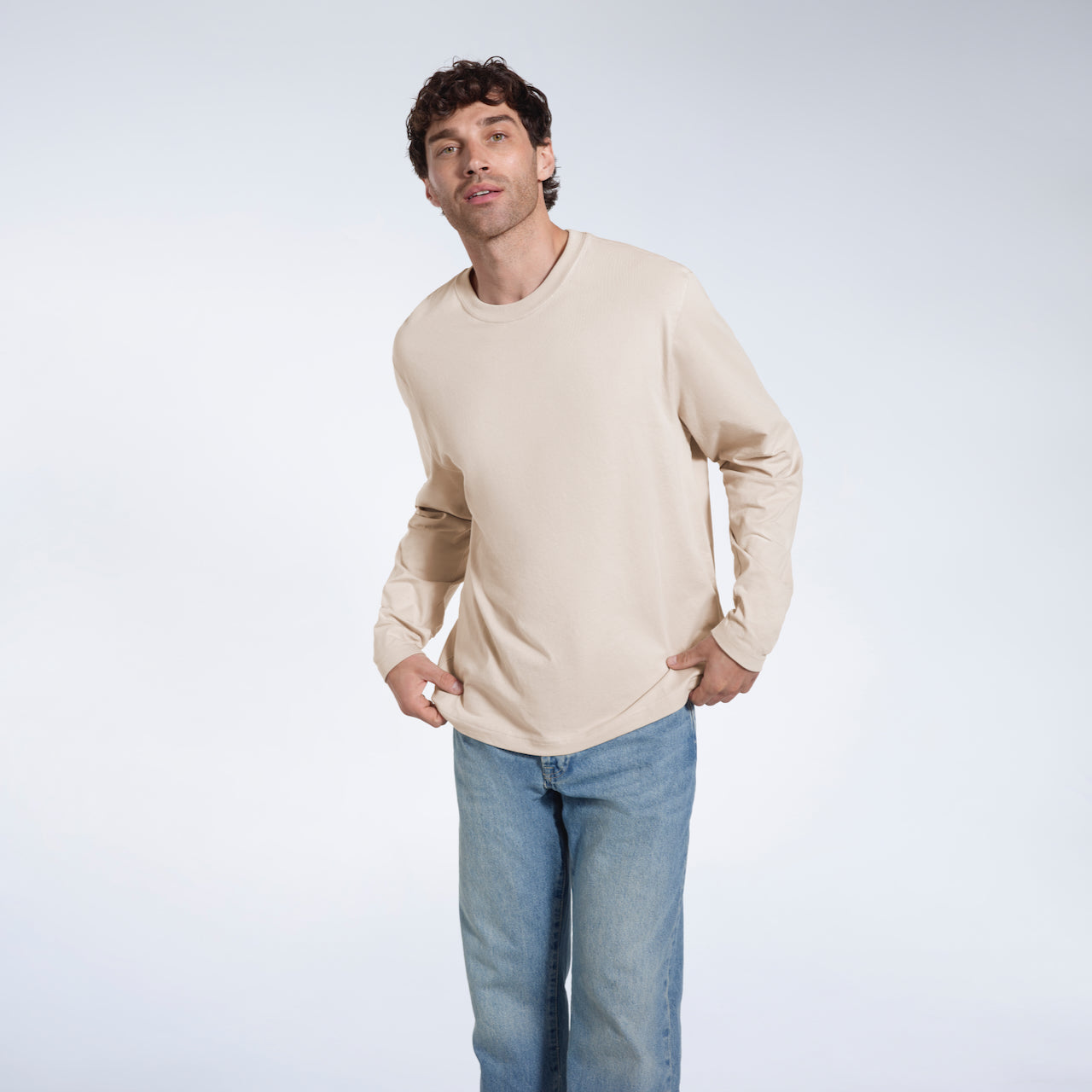 A man looking into the camera, touching the bottom of his plain stone long sleeve organic cotton t shirt.