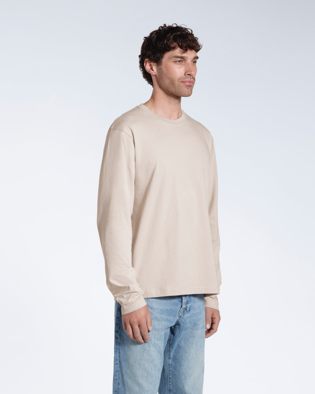 A man looking away from the camera with his arms by his sides, modelling a plain stone long sleeve organic cotton t shirt.