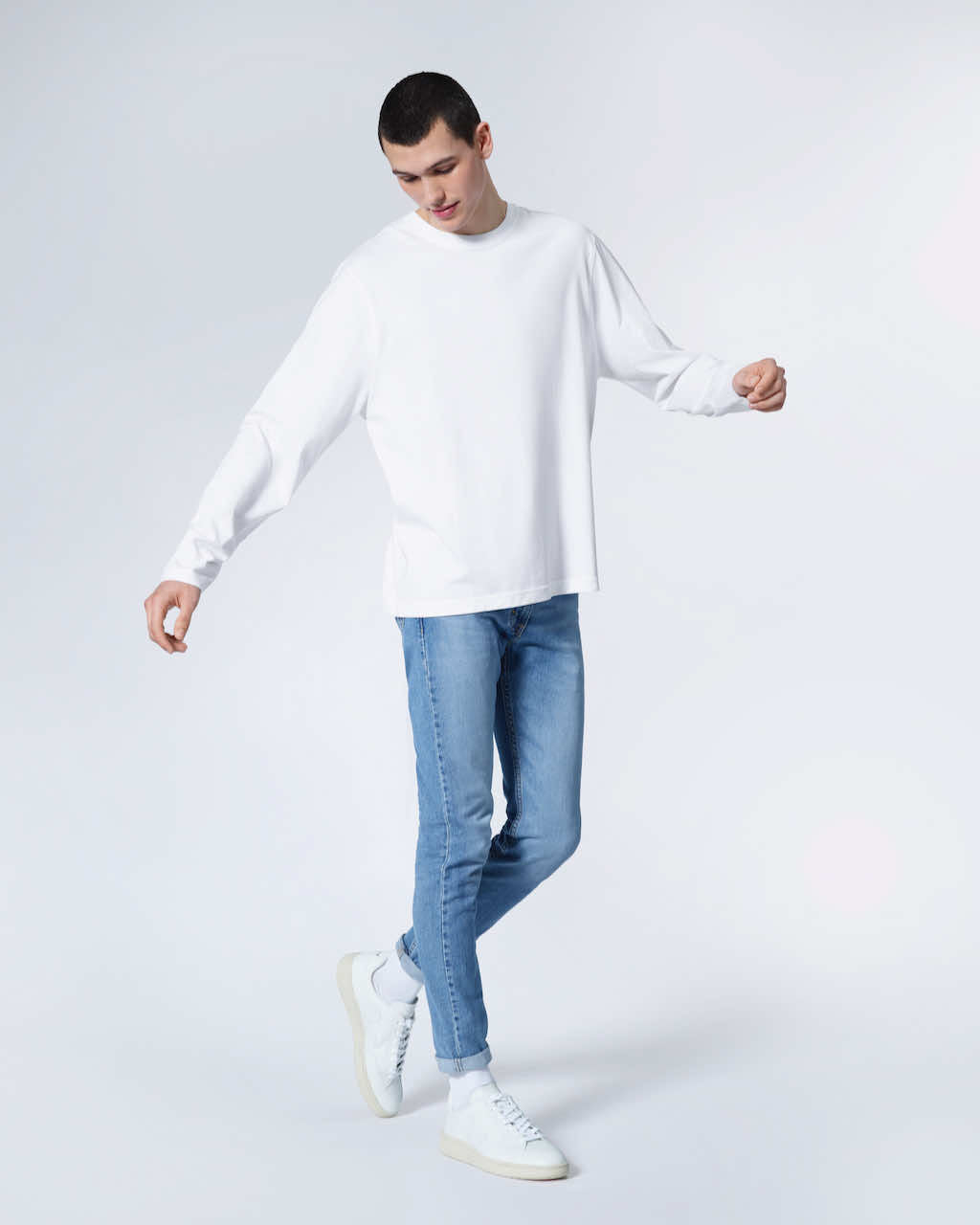A full body photo of a man looking down with his arms out, modelling a plain white long sleeve organic cotton t shirt.