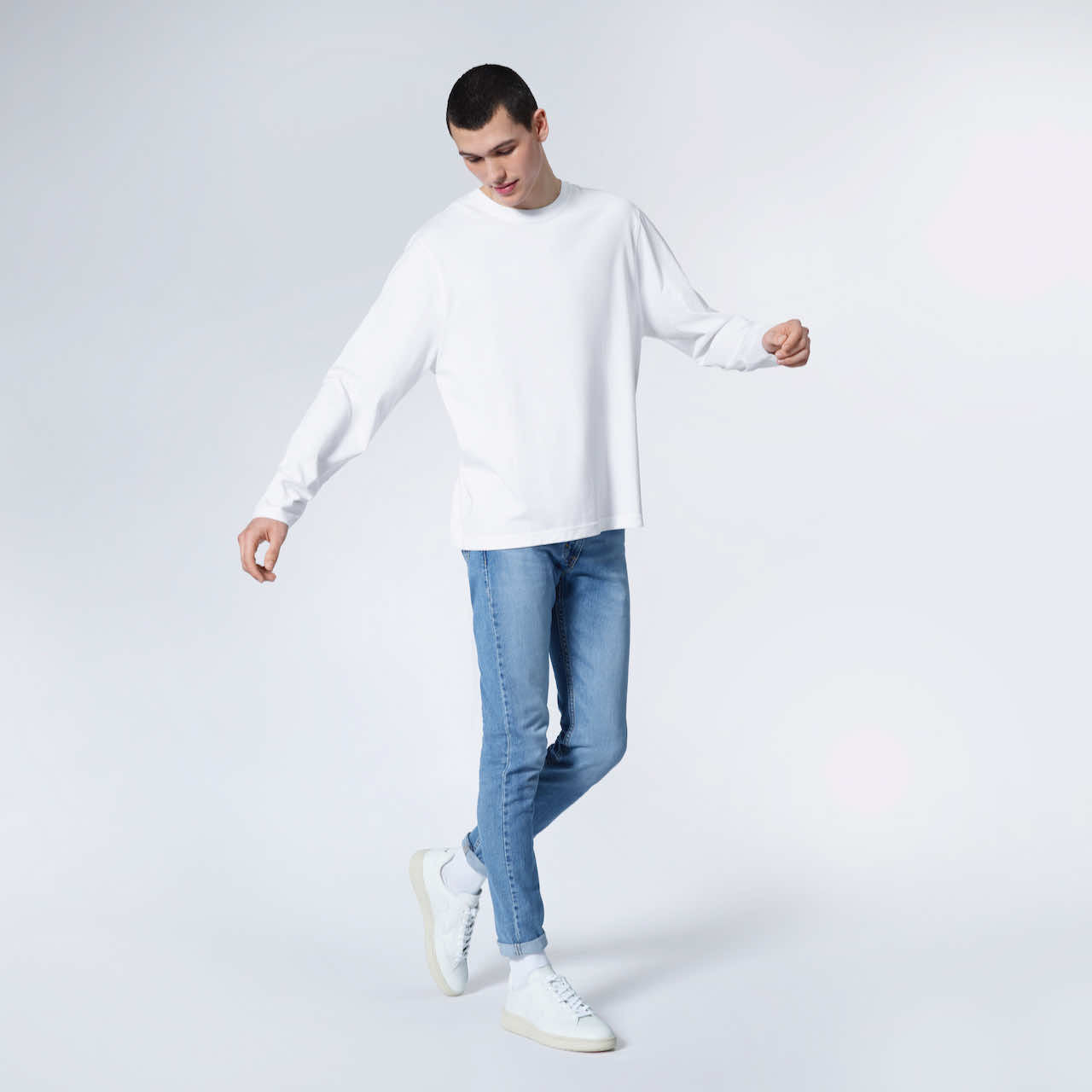A full body photo of a man looking down with his arms out, modelling a plain white long sleeve organic cotton t shirt.