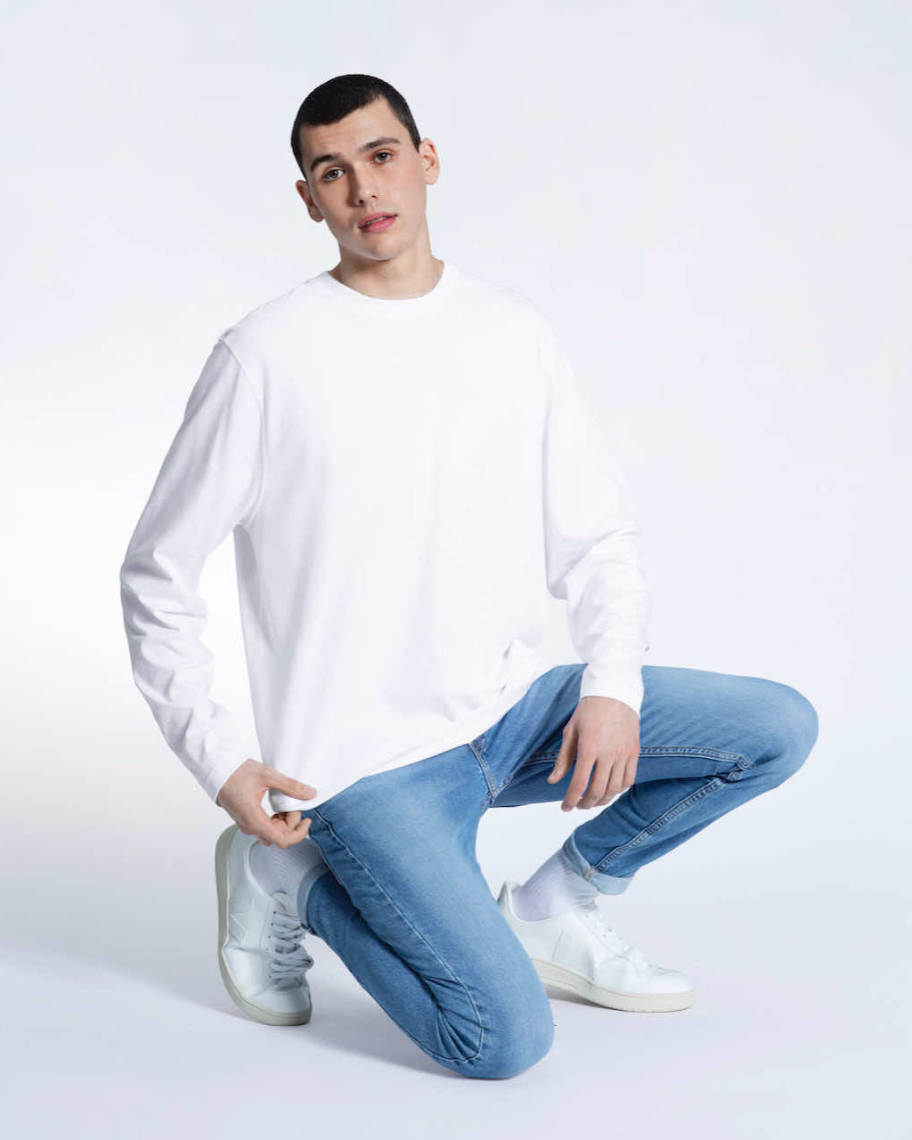 A man kneeling down looking into the camera, modelling a plain white long sleeve organic cotton t shirt.