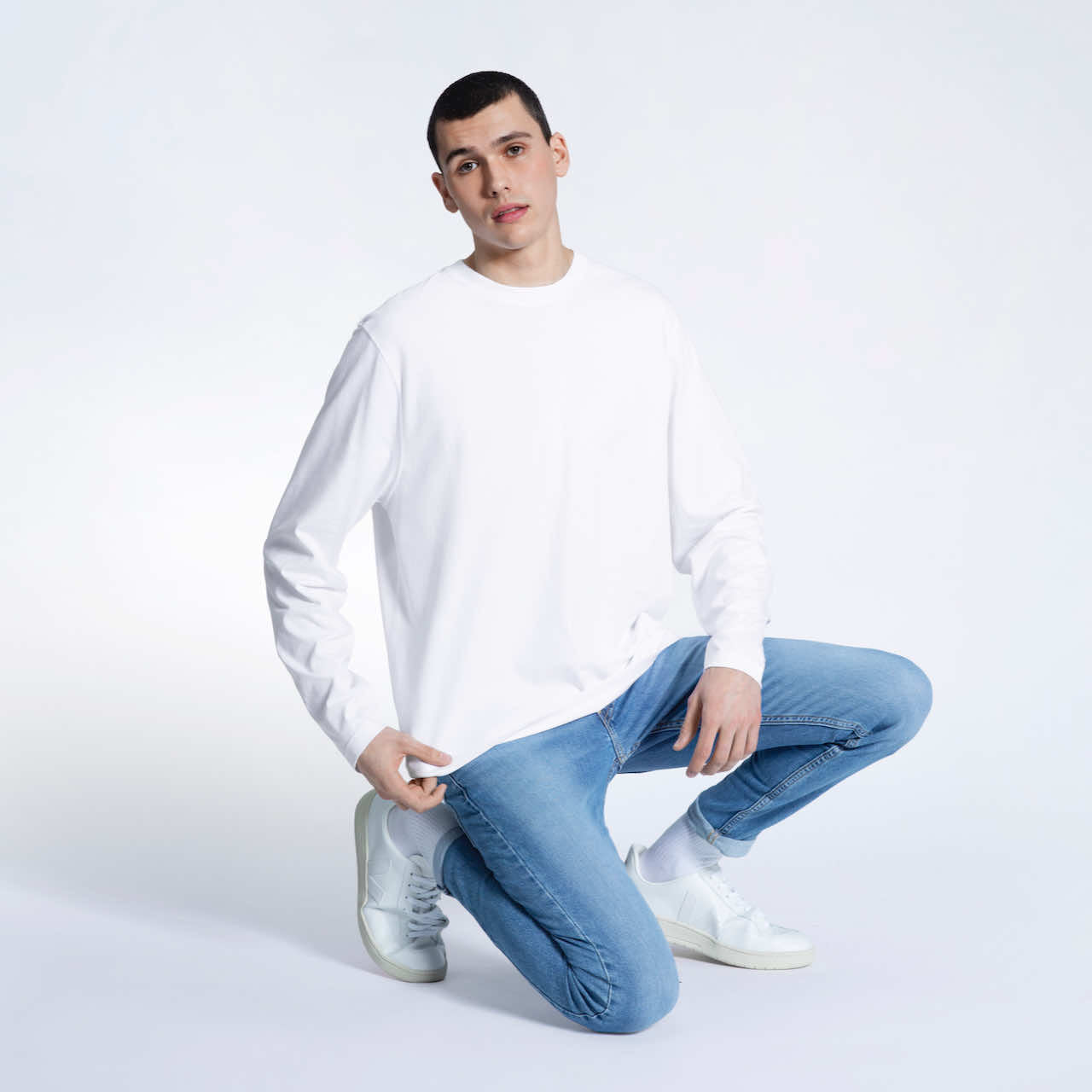 A man kneeling down looking into the camera, modelling a plain white long sleeve organic cotton t shirt.