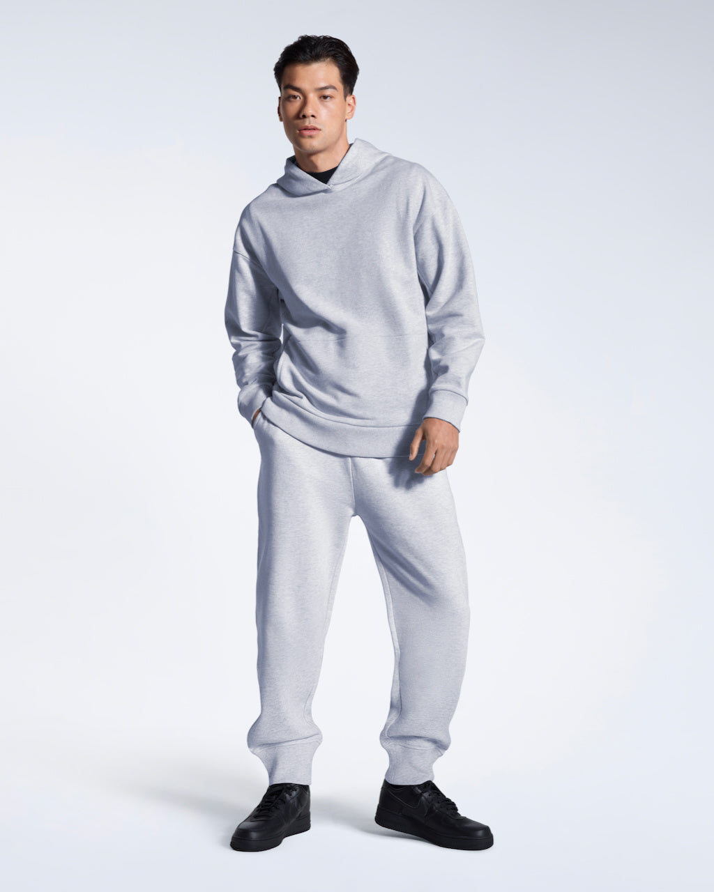 A man looking into the camera with a hand in his pocket, modelling a pair of plain grey, organic cotton joggers with a matching oversized hoodie.