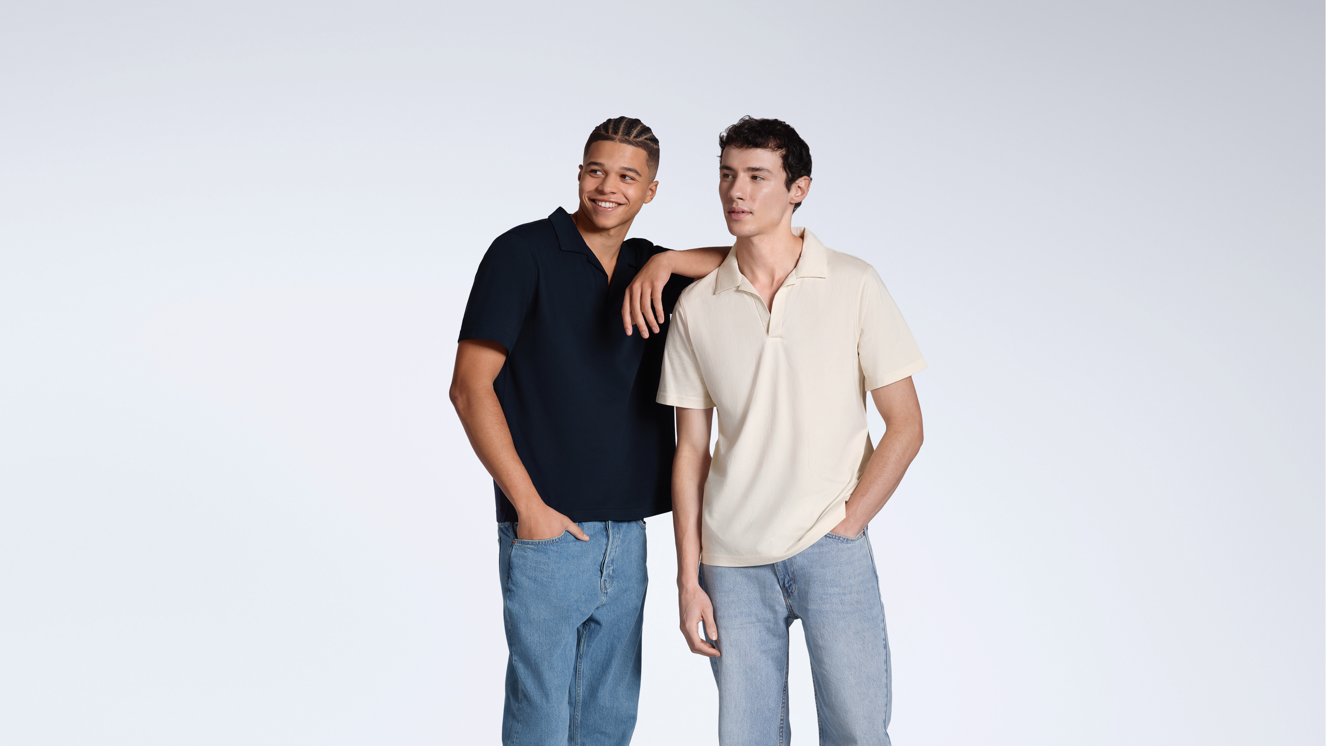 Two men looking away from the camera, modelling plain navy and natural, organic cotton polo shirts.