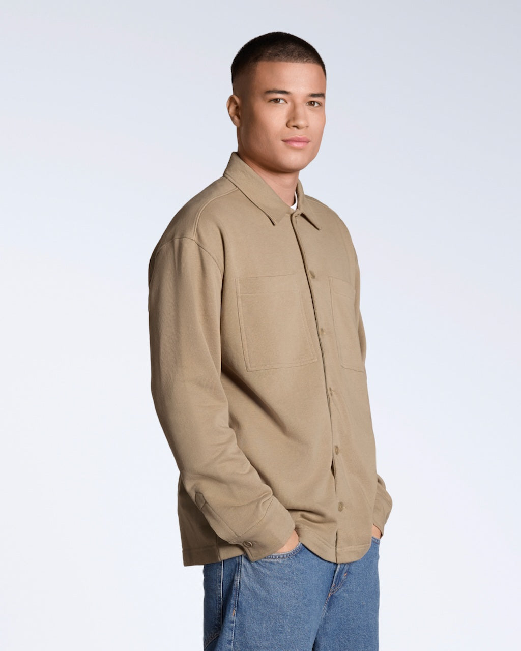 A man looking into the camera with his hands in his pockets, modelling a plain beige, organic cotton overshirt.