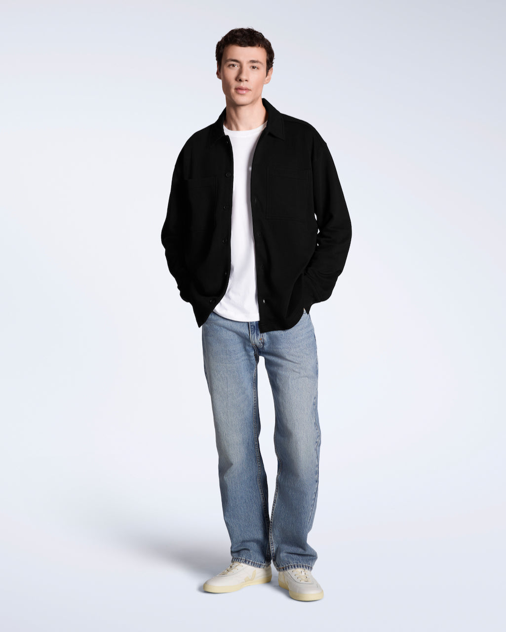 A full body photo of a man looking into the camera with his hands in his pockets, modelling a plain black, organic cotton overtshirt with a pair of jeans.