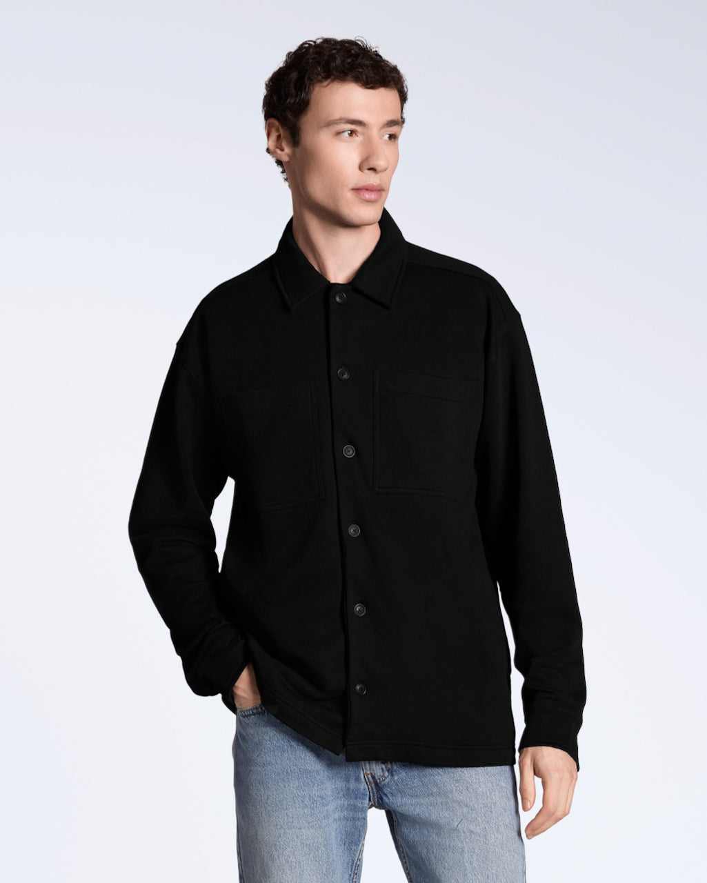 A man looking away from the camera with his hand in his pocket, modelling a plain black, organic cotton overshirt.