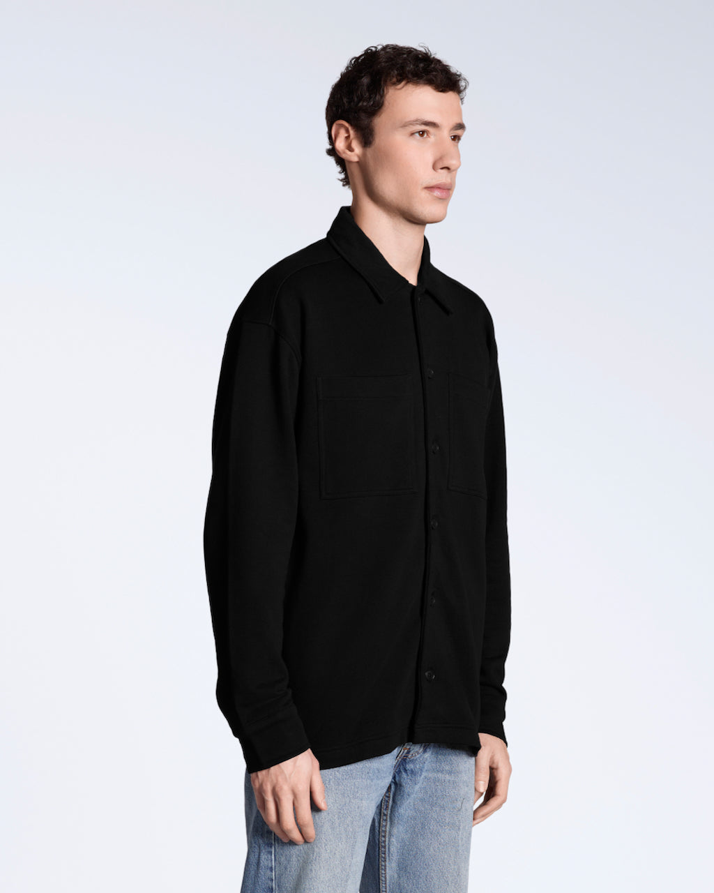 A man at an angle with his arms by his sides, modelling a plain black, organic cotton overshirt.