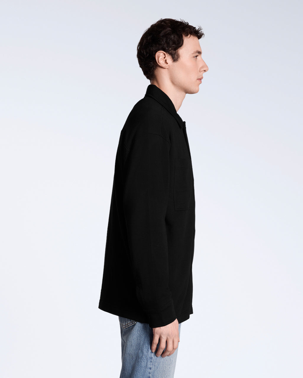 A man facing side on, modelling a plain black, organic cotton overshirt.
