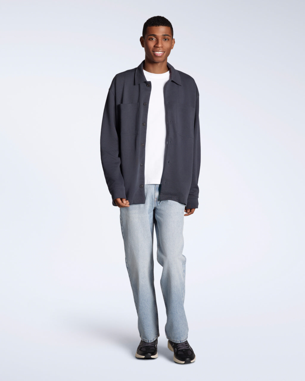 A full body photo of a man smiling into the camera, modelling a plain dark grey, organic cotton overtshirt with a pair of jeans.