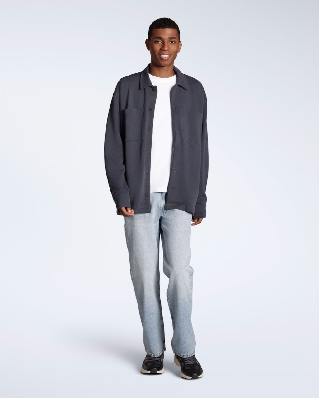 A full body photo of a man smiling into the camera, modelling a plain dark grey, organic cotton overtshirt with a pair of jeans.