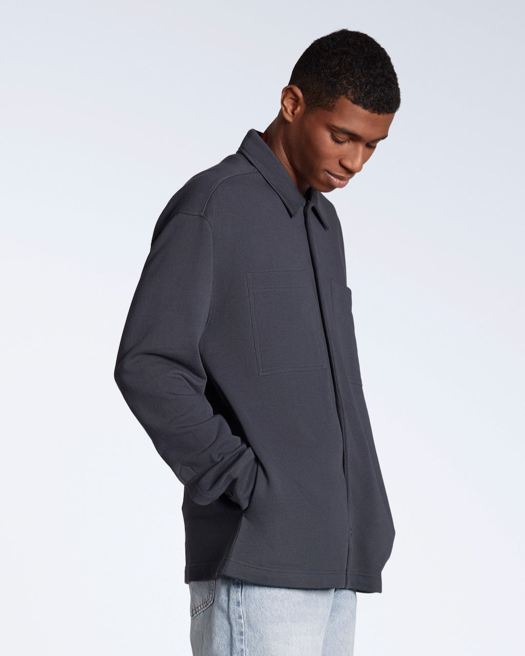 A man looking down with his hands in his pockets, modelling a plain dark grey, organic cotton overshirt.