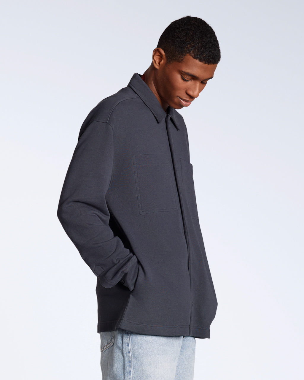 A man looking down with his hands in his pockets, modelling a plain dark grey, organic cotton overshirt.