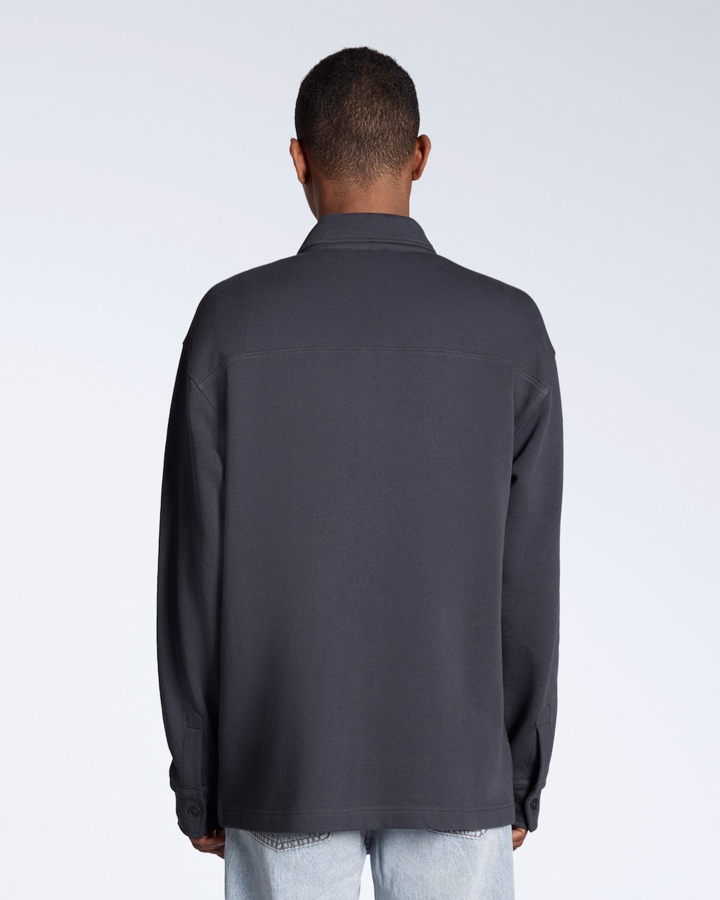 A man facing away from the camera, modelling a plain dark grey, organic cotton overshirt.