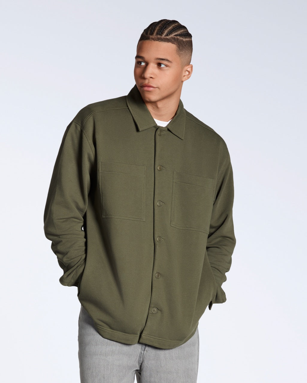 A man looking away from the camera with his hands in his pockets, modelling a plain olive coloured, organic cotton overshirt.
