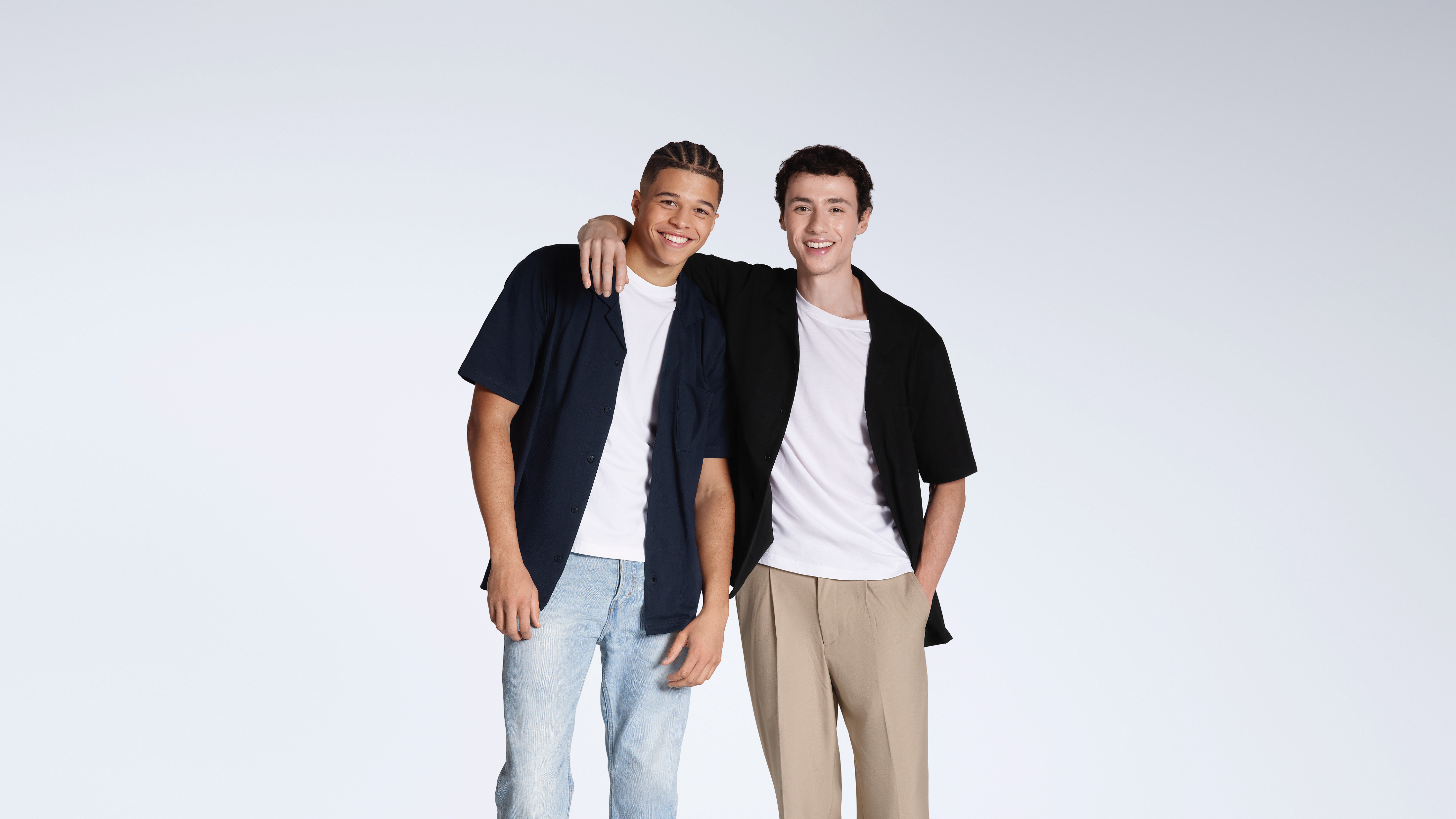 Two men smiling into the camera with their arms round eachother, modelling plain black and navy, organic cotton resort shirts.