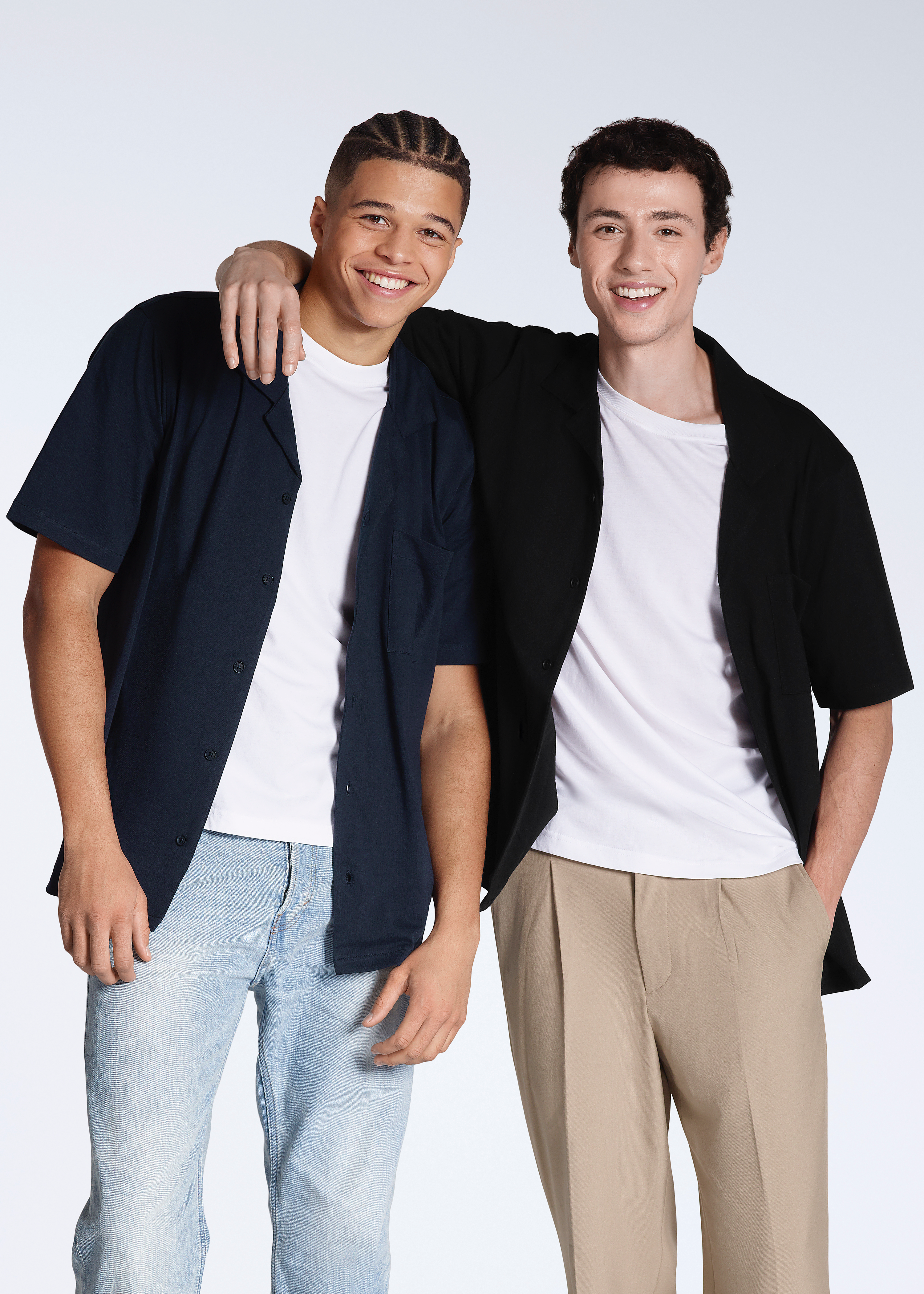 Two men smiling into the camera with their arms round eachother, modelling plain black and navy, organic cotton resort shirts.
