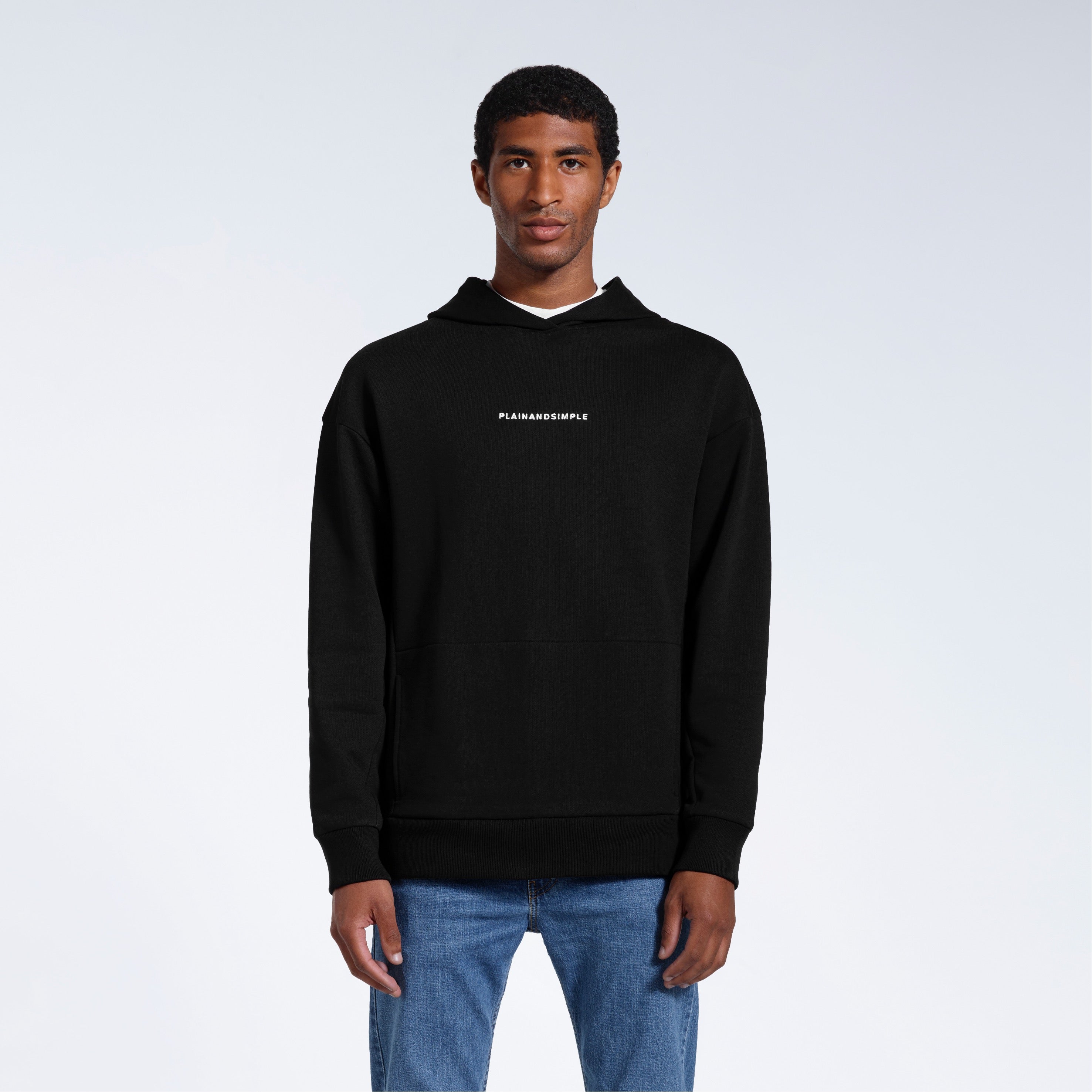 A man facing front on looking into the camera with his arms by his sides, modelling a branded black oversized heavyweight organic cotton hoodie.