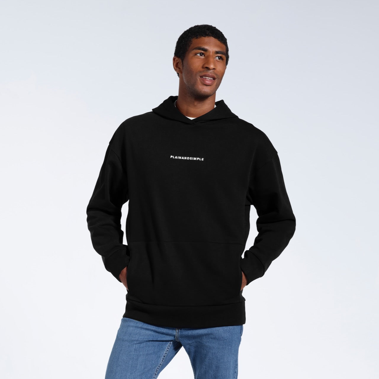 A man with his hands in his pockets looking away from the camera, modelling a branded black oversized heavyweight organic cotton hoodie.