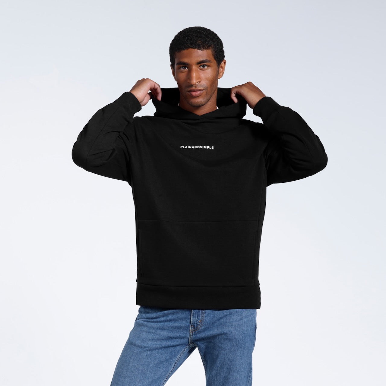 A man touching his hood and looking into the camera, modelling a branded black oversized heavyweight organic cotton hoodie.