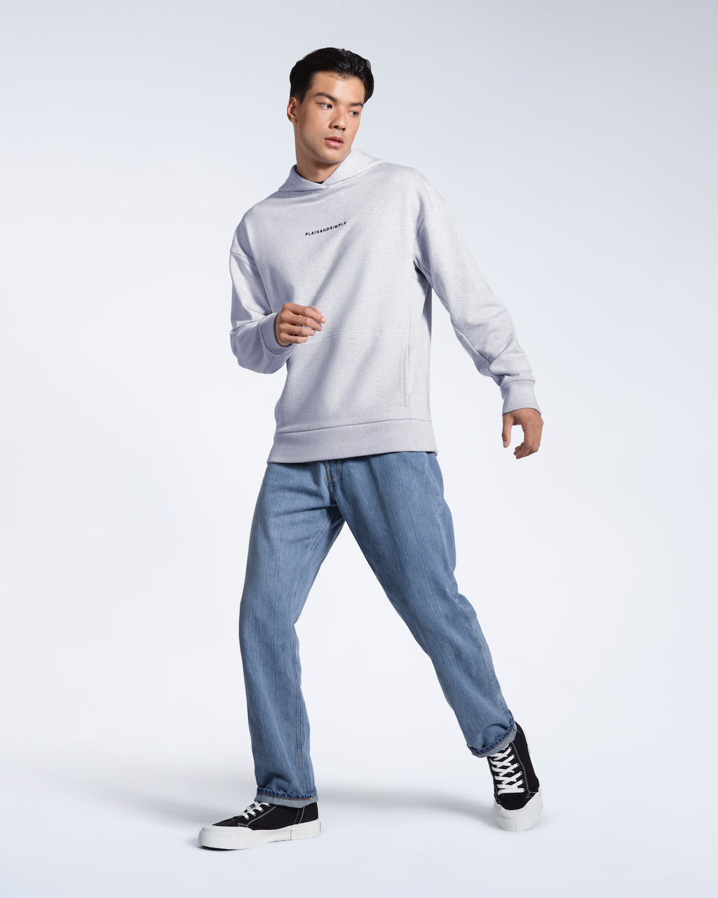 A full body photo of a man walking looking away from the camera, modelling a grey branded heavyweight organic cotton hoodie.