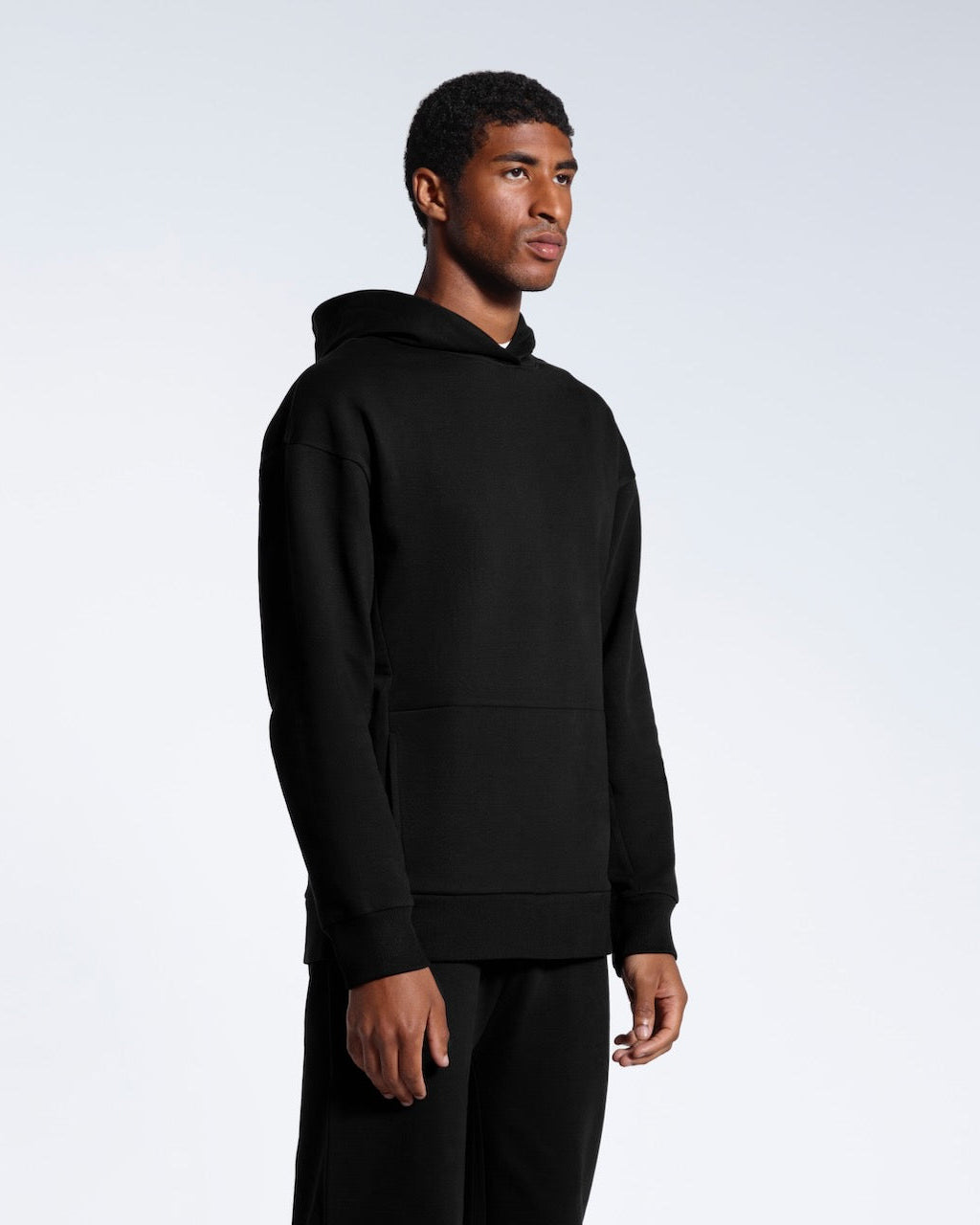 A man at an angle with his arms by his sides, modelling a black oversized heavyweight organic cotton hoodie with matching joggers. 