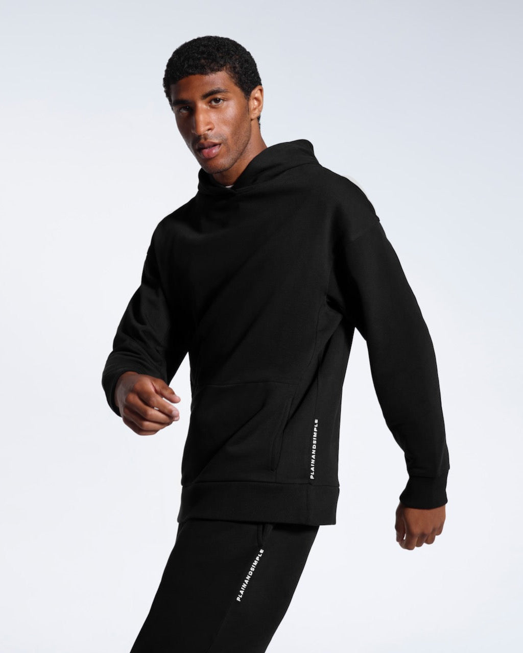 A man mid movement looking into the camera, modelling a black branded oversized heavyweight organic cotton hoodie with matching joggers. 