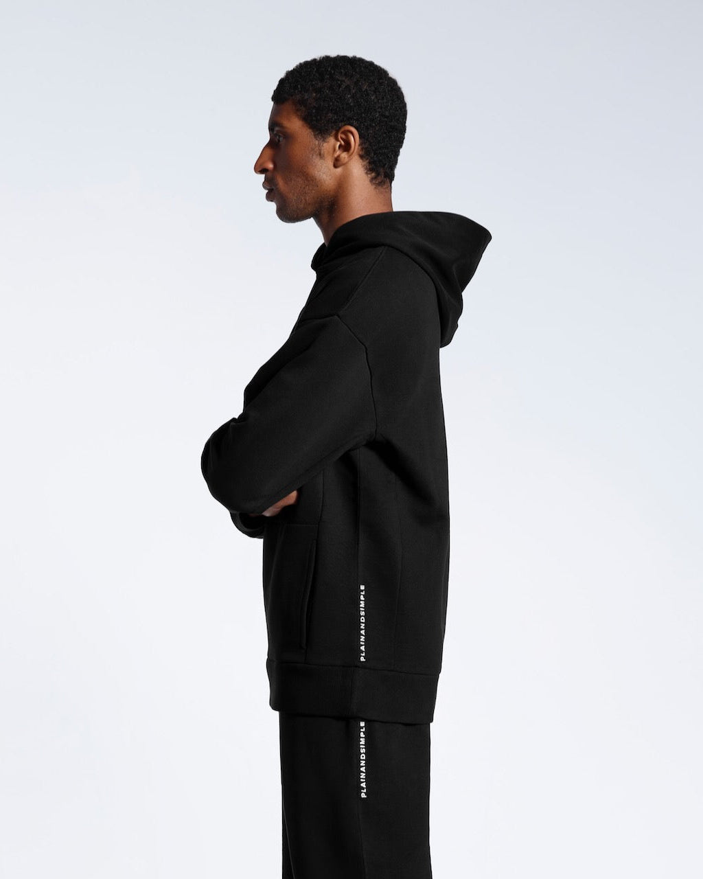 A man facing side on with his arms crossed, modelling a black branded oversized heavyweight organic cotton hoodie with matching joggers. 
