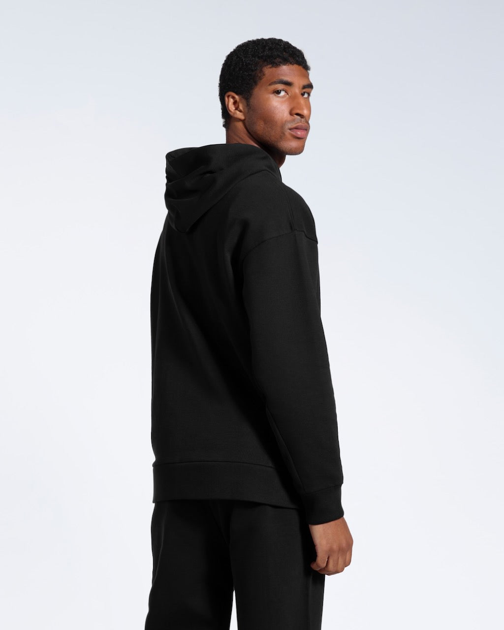 A man facing away from the camera looking back, modelling a black oversized heavyweight organic cotton hoodie with matching joggers. 