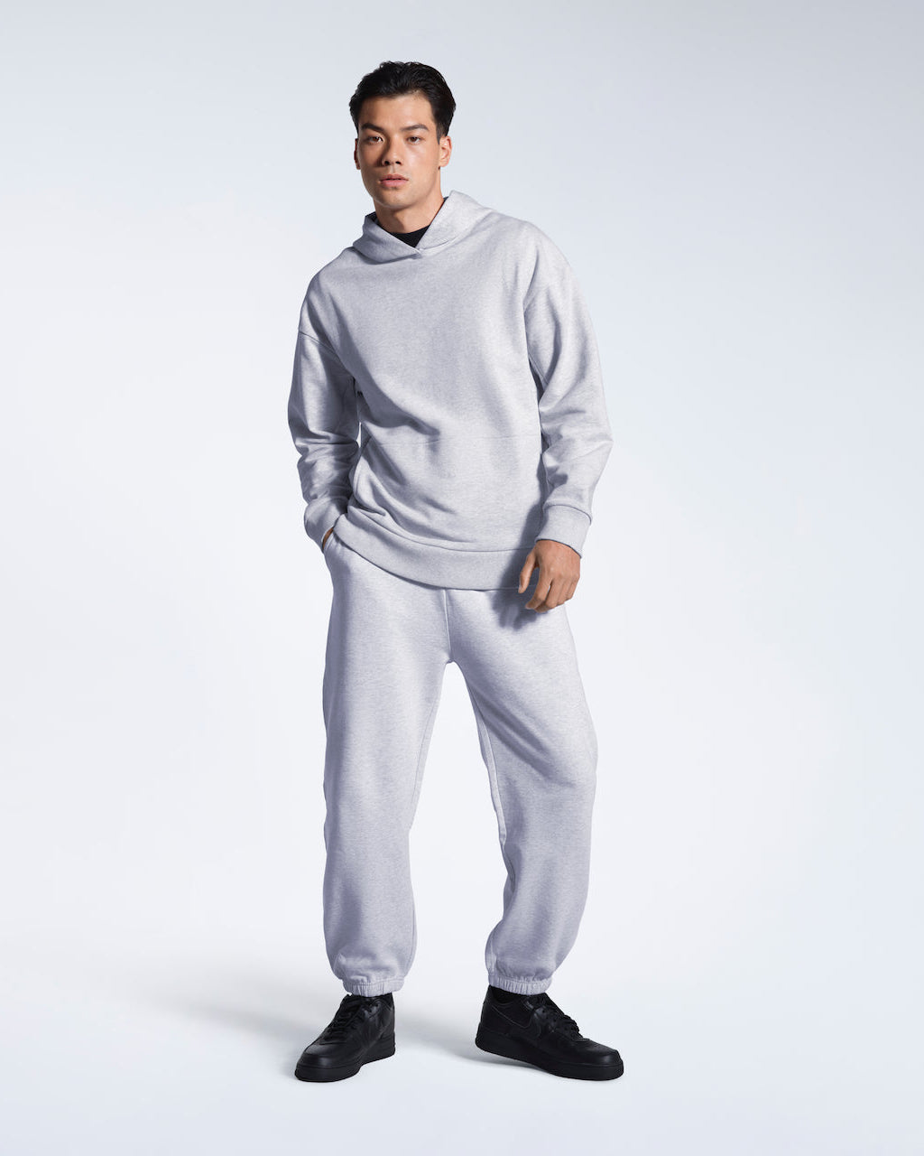 A full body photo of a man looking into the camera with a hand in his pocket, modelling a grey oversized heavyweight organic cotton hoodie with matching joggers.