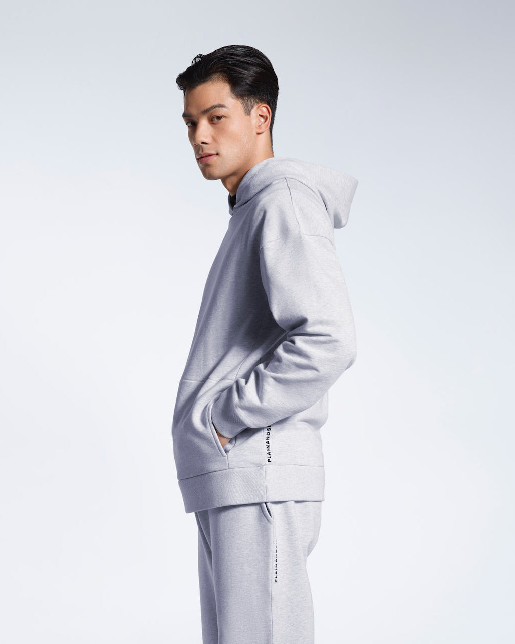 A man facing side on looking into the camera, modelling a branded grey oversized heavyweight organic cotton hoodie with matching joggers.