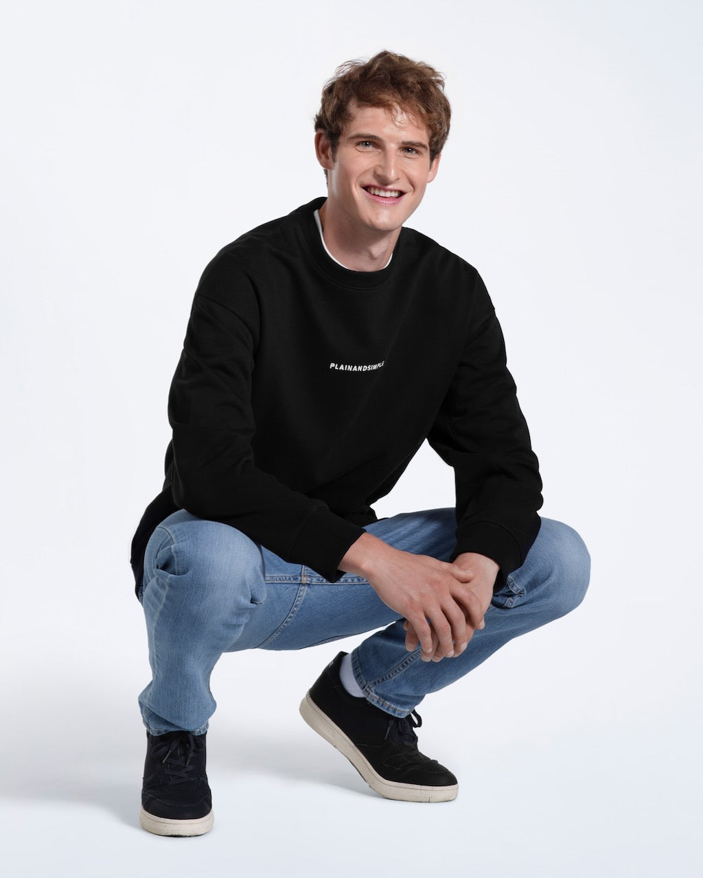 A man crouching down smiling, modelling a branded black oversized heavyweight organic cotton sweatshirt.