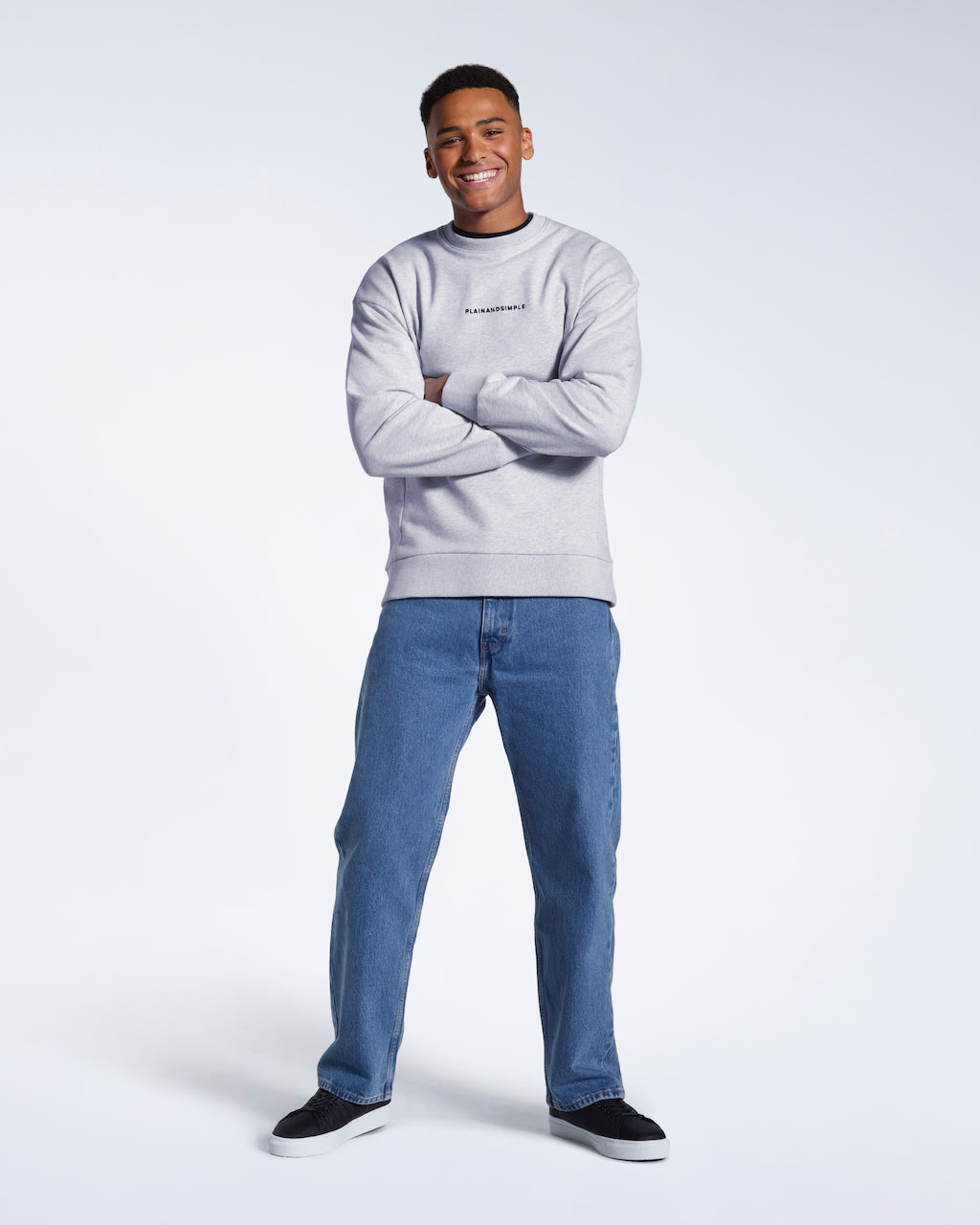 A full body photo of a man smiling into the camera with his arms folded, modelling a branded grey oversized heavyweight organic cotton sweatshirt.