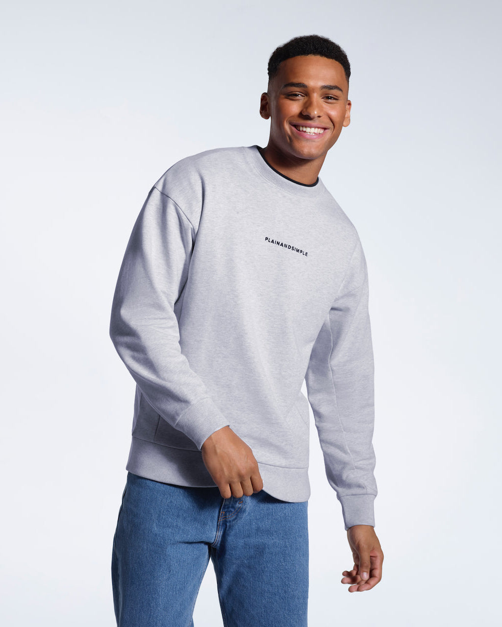 A man smiling into the camera, modelling a grey branded oversized heavyweight organic cotton sweatshirt.