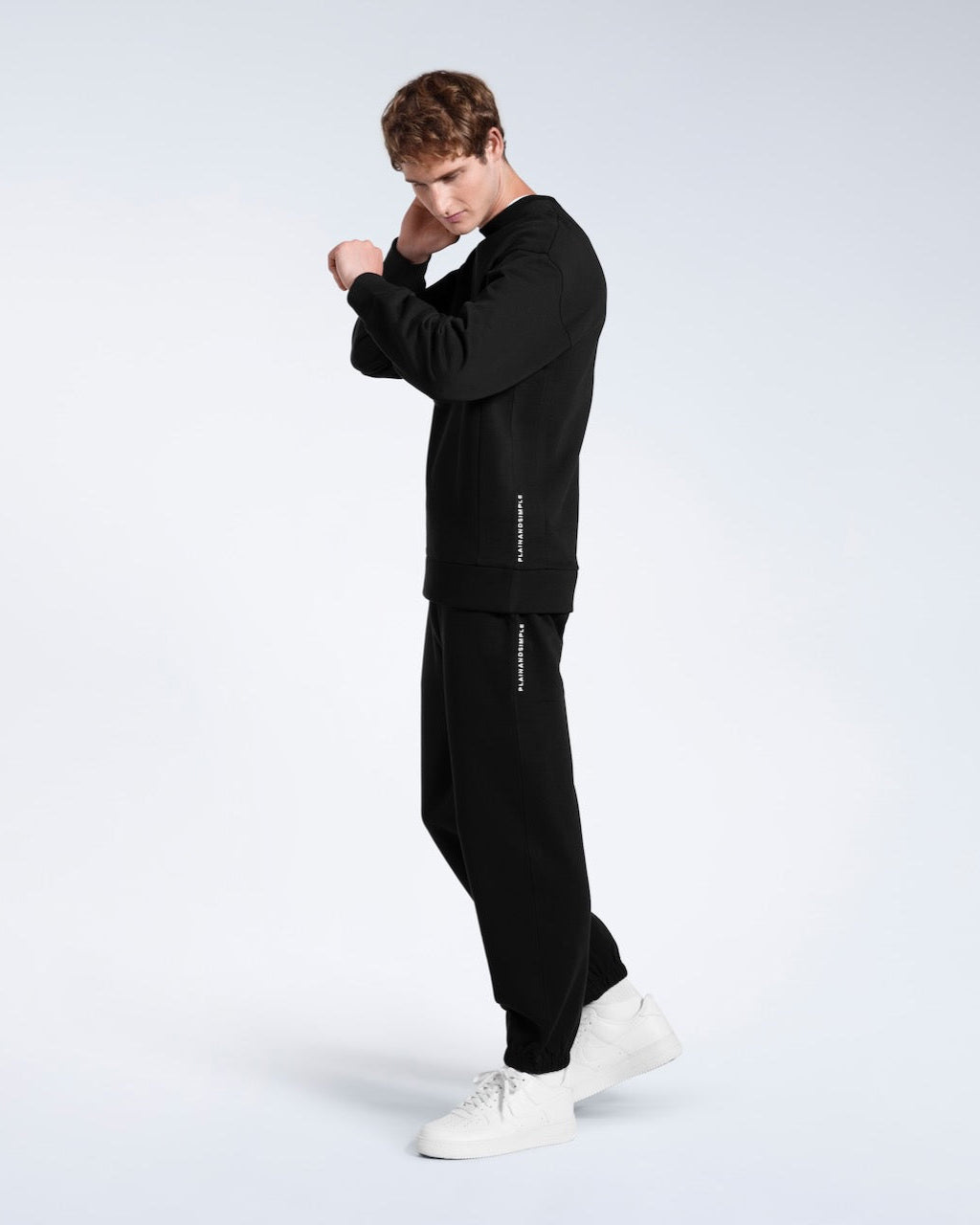 A full body photo of a man with his arms raised, modelling a branded black oversized heavyweight organic cotton sweatshirt with matching joggers.