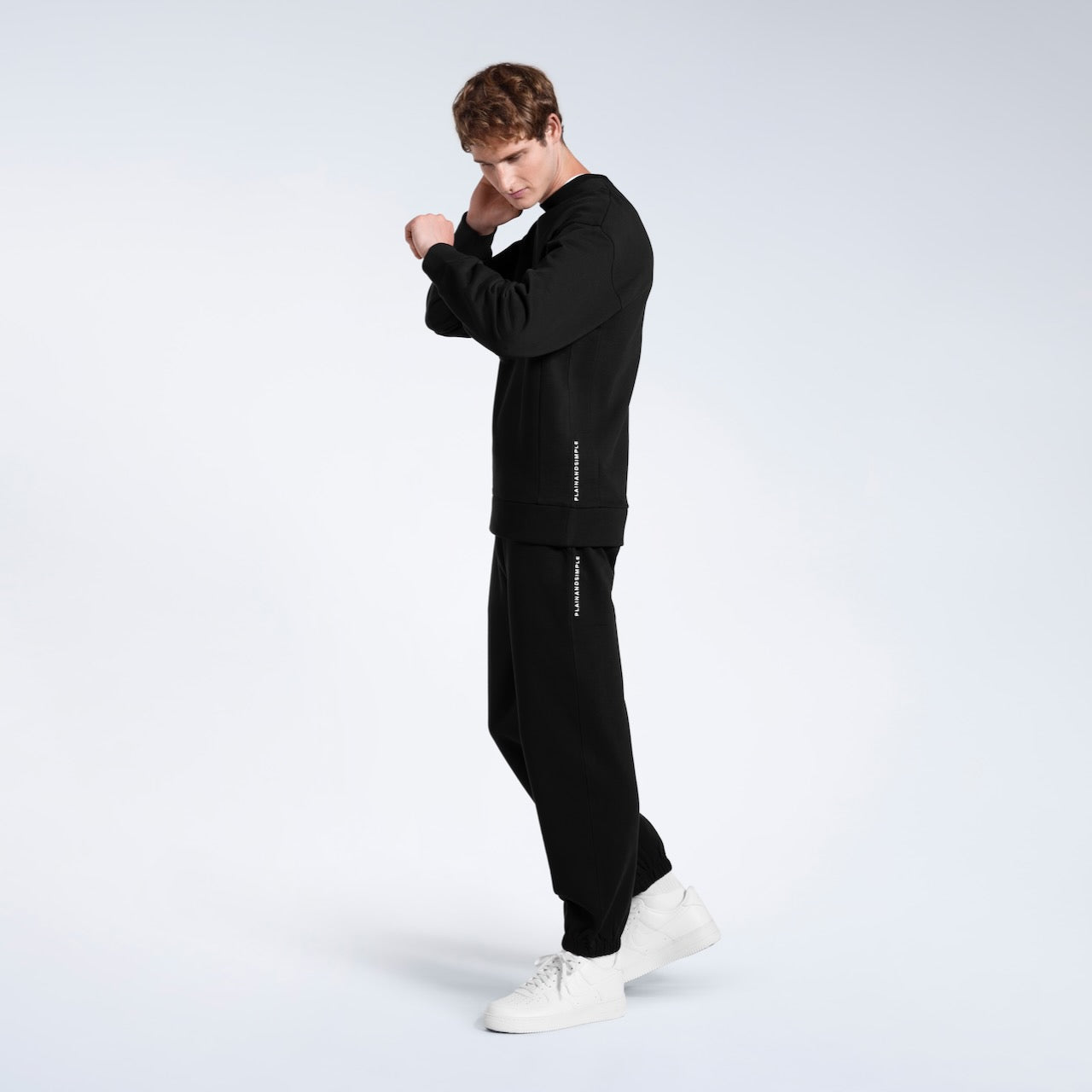 A full body photo of a man with his arms raised, modelling a branded black oversized heavyweight organic cotton sweatshirt with matching joggers.