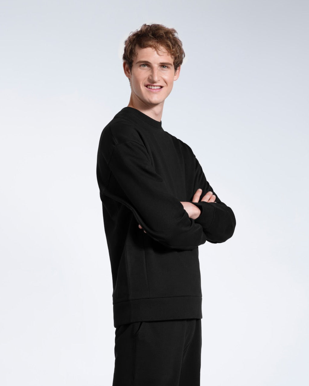 A man smiling into the camera with his arms folded, modelling a black heavyweight organic cotton sweatshirt with matching joggers.