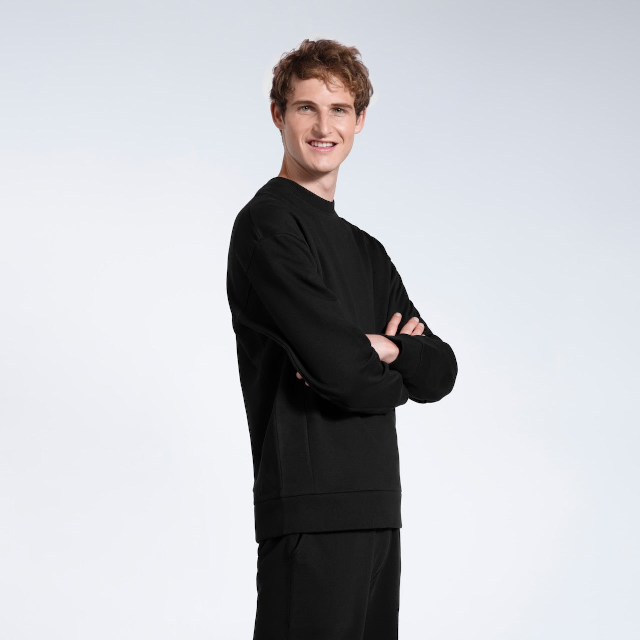A man smiling into the camera with his arms folded, modelling a black heavyweight organic cotton sweatshirt with matching joggers.