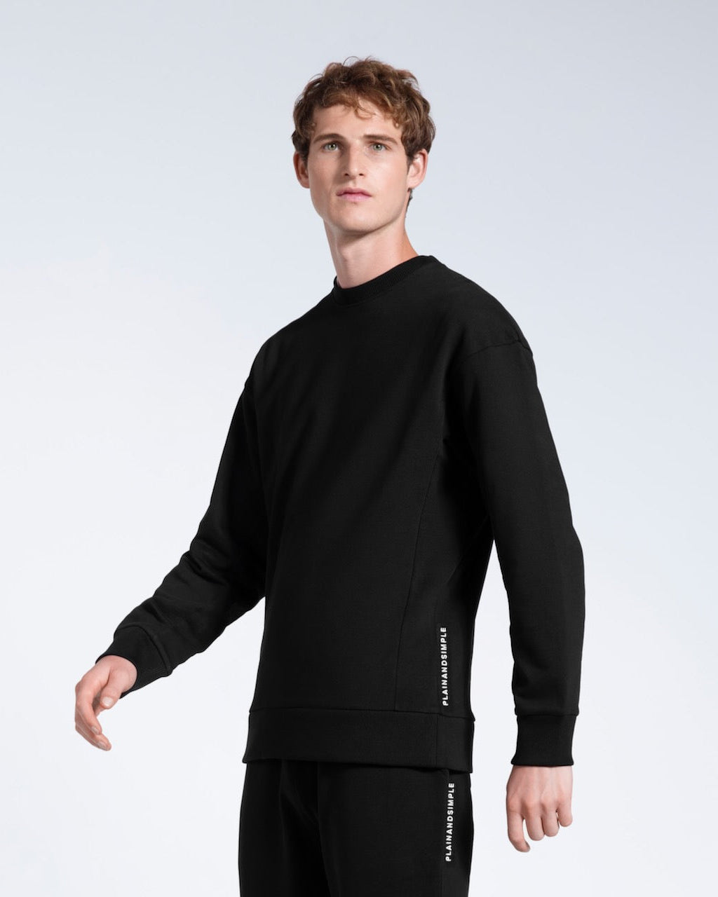 A man looking into the camera, modelling a branded black heavyweight organic cotton sweatshirt with matching joggers.
