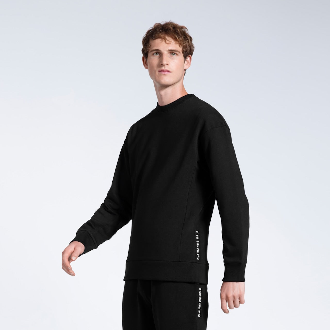 A man looking into the camera, modelling a branded black heavyweight organic cotton sweatshirt with matching joggers.