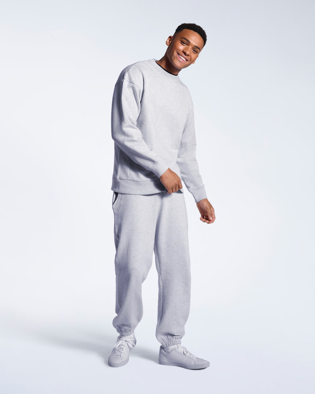 A full body photo of a man smiling into the camera, modelling a grey oversized heavyweight organic cotton sweatshirt with matching joggers.